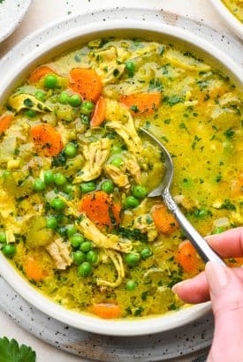A large shallow bowl of bright yellow turmeric chicken soup, brimming with veggies and fresh herbs, a spoon dipping into the bowl.