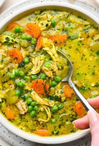 A large shallow bowl of bright yellow turmeric chicken soup, brimming with veggies and fresh herbs, a spoon dipping into the bowl.