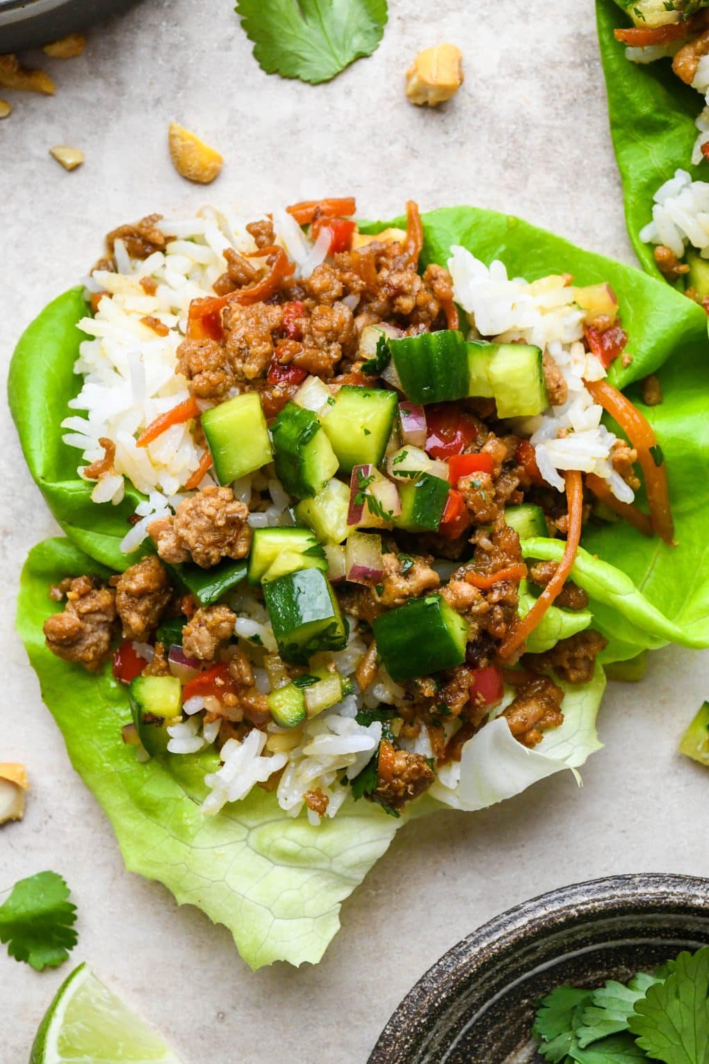 A single lettuce wrap with white rice on a cream colored background, topped with diced asian cucumber salad, chopped cashews, fresh cilantro, and lime wedges.