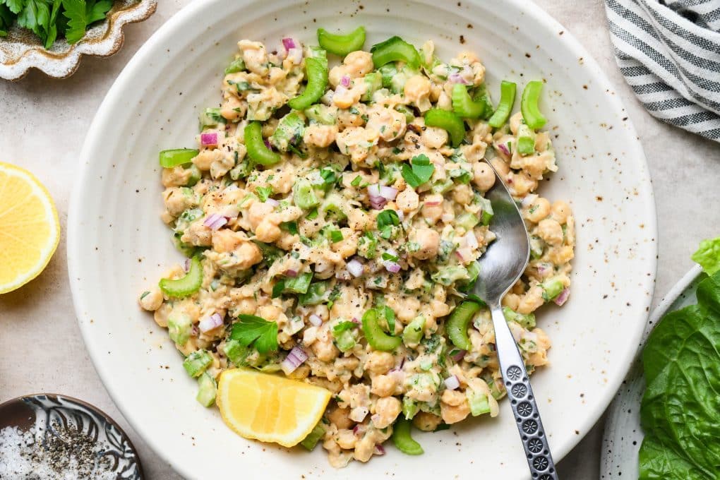 How to make smashed chickpea salad: Chickpea salad in a large shallow bowl with fresh herbs a lemon wedge, and a serving spoon angled into the bowl. On a light brown background, surrounded by various ingredients and a striped napkin.
