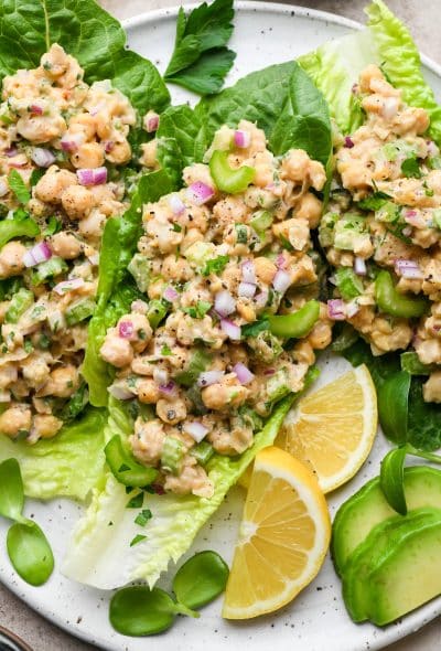 Smashed chickpea salad on romaine lettuce leaves, on a large plate surrounded by lemon wedges, sliced avocado, and fresh herbs.