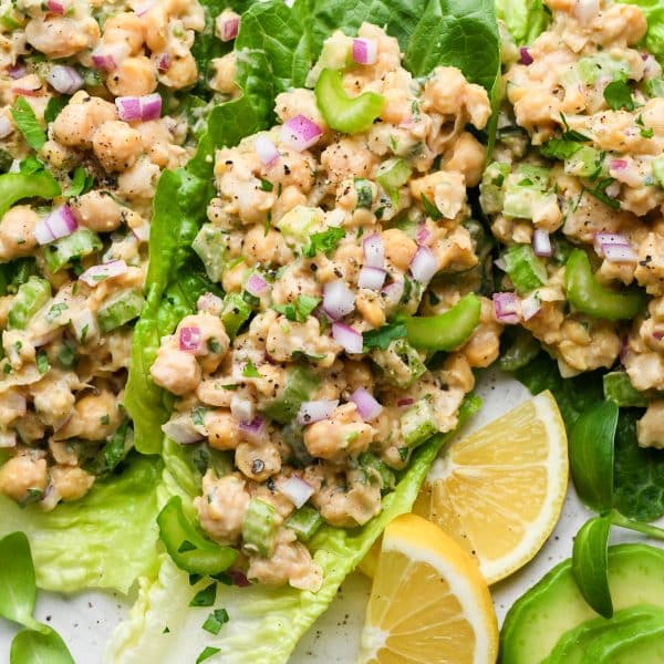 Smashed chickpea salad on romaine lettuce leaves, on a large plate surrounded by lemon wedges, sliced avocado, and fresh herbs.