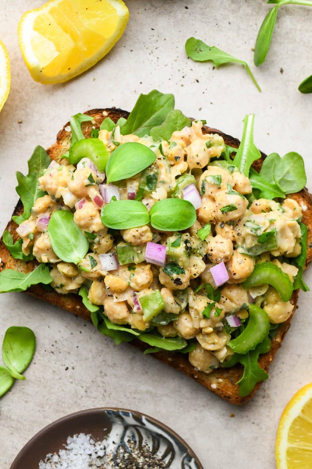 Smashed chickpea salad on a piece of bread for a sandwich, topped with fresh sprouts on a light brown textured background.