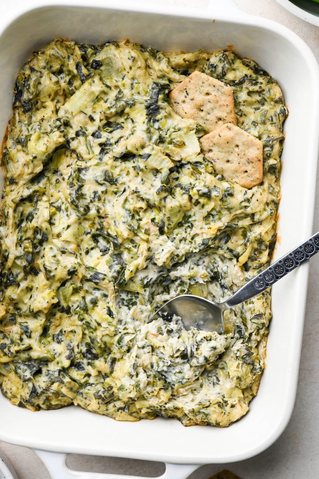 Baked dairy free spinach artichoke dip in a small rectangular baking dish with some crackers tucked into the dip and a spoon scooping some out.