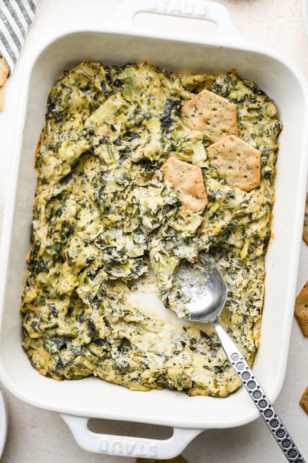 A small cream colored baking dish of baked dairy free spinach artichoke dip, with a spoon angled into the dish where some dip has been scooped out.