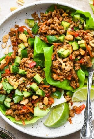 A shallow white speckled bowl filled with two asian style pork lettuce wraps, topped with diced cucumber salad alongside some lime wedges.