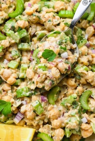 A spoon lifting out a spoonful of smashed chickpea salad of a bowl full of the salad.