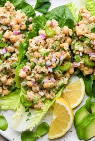 Smashed chickpea salad on romaine lettuce leaves, on a large plate surrounded by lemon wedges, sliced avocado, and fresh herbs.