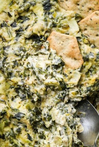 Close up image of a cracker being dipped into creamy dairy free spinach artichoke dip.