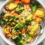 A wide ceramic bowl filled with brown rice, colorful roasted vegetables, ground chicken patties, and topped with a bright yellow turmeric tahini sauce and fresh herbs.
