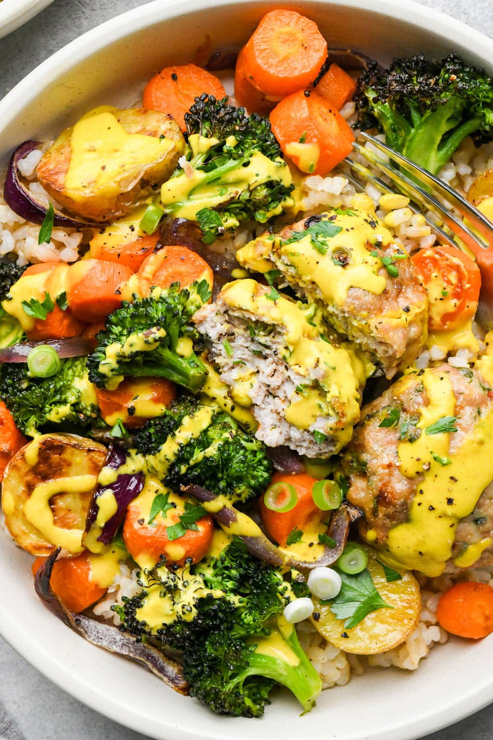 A wide ceramic bowl filled with brown rice, colorful roasted vegetables, ground chicken patties cut in half, topped with a bright yellow turmeric tahini sauce and fresh herbs.