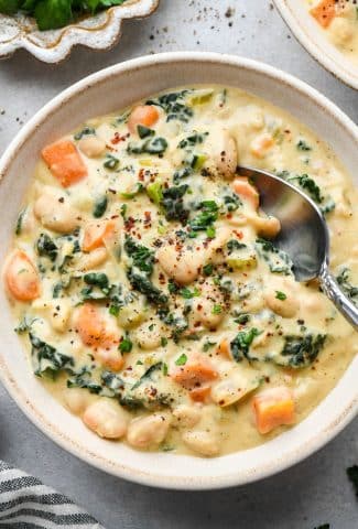 A creamy ceramic bowl of creamy white bean and kale soup with a soup spoon dipping into the bowl for a bite.