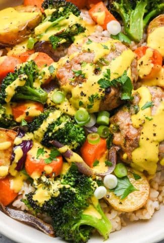 Close up image of a wide ceramic bowl filled with brown rice, colorful roasted vegetables, ground chicken patties, and topped with a bright yellow turmeric tahini sauce and fresh herbs.