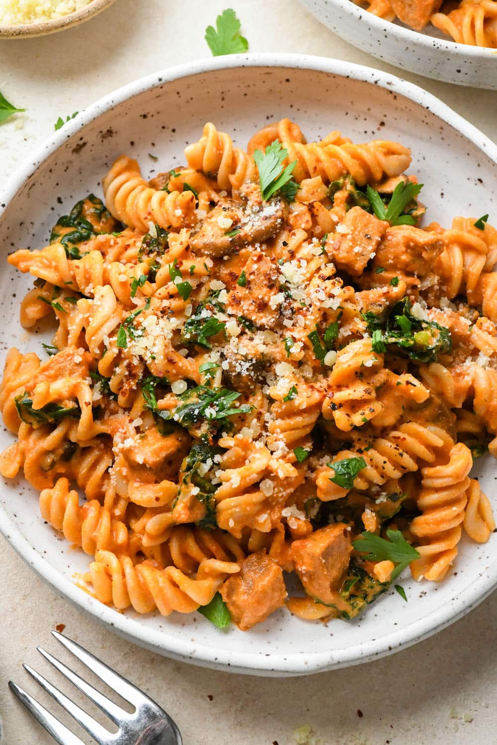 Creamy tomato sausage pasta in a shallow white speckled bowl, next to another bowl of pasta on a light cream background, and topped with dairy free parmesan, chili flakes, and a fresh herbs.