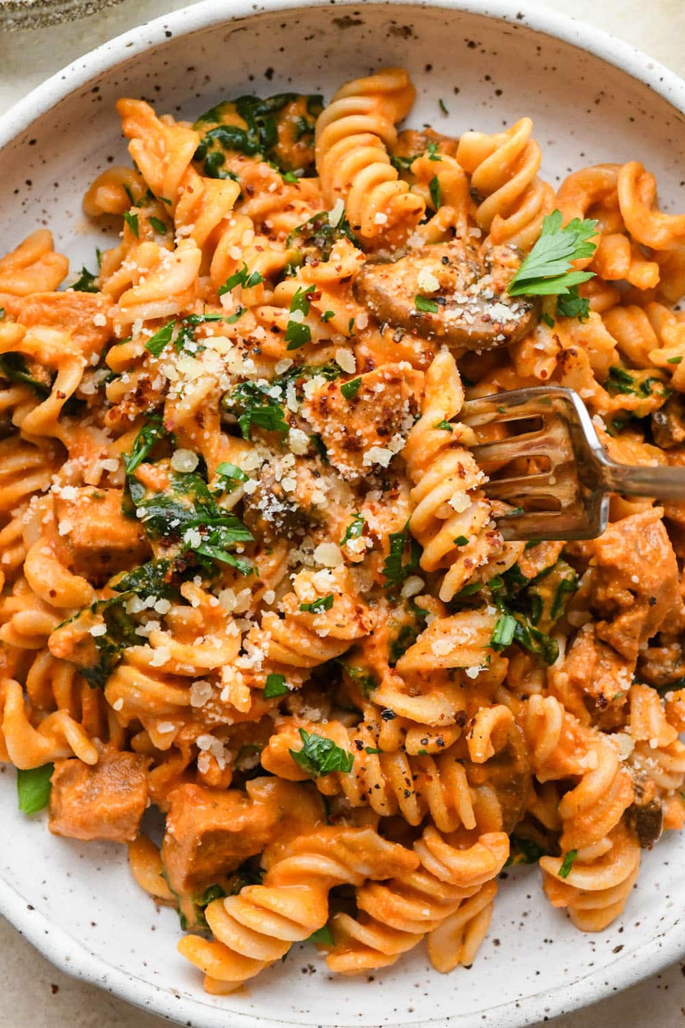Creamy tomato sausage pasta in a shallow white speckled bowl, topped with dairy free parmesan, chili flakes, and a fresh herbs, with a fork lifting out a bite of pasta.