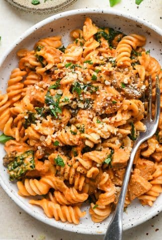 Creamy tomato sausage pasta in a shallow white speckled bowl, topped with dairy free parmesan, chili flakes, and a fresh herbs.