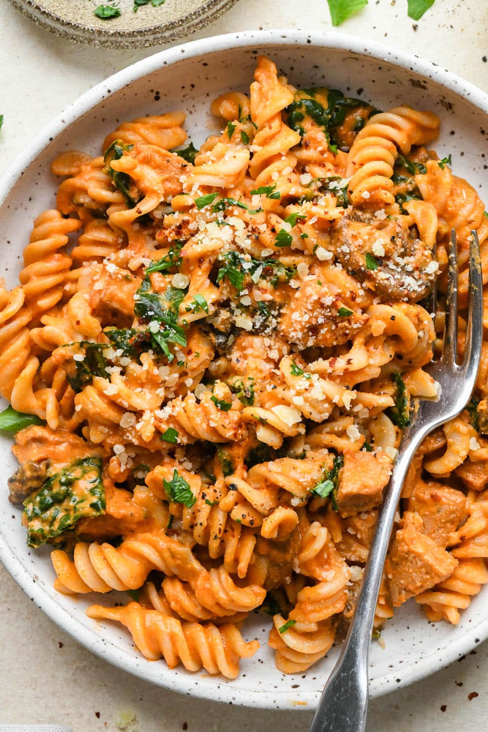 Creamy tomato sausage pasta in a shallow white speckled bowl, topped with dairy free parmesan, chili flakes, and a fresh herbs.