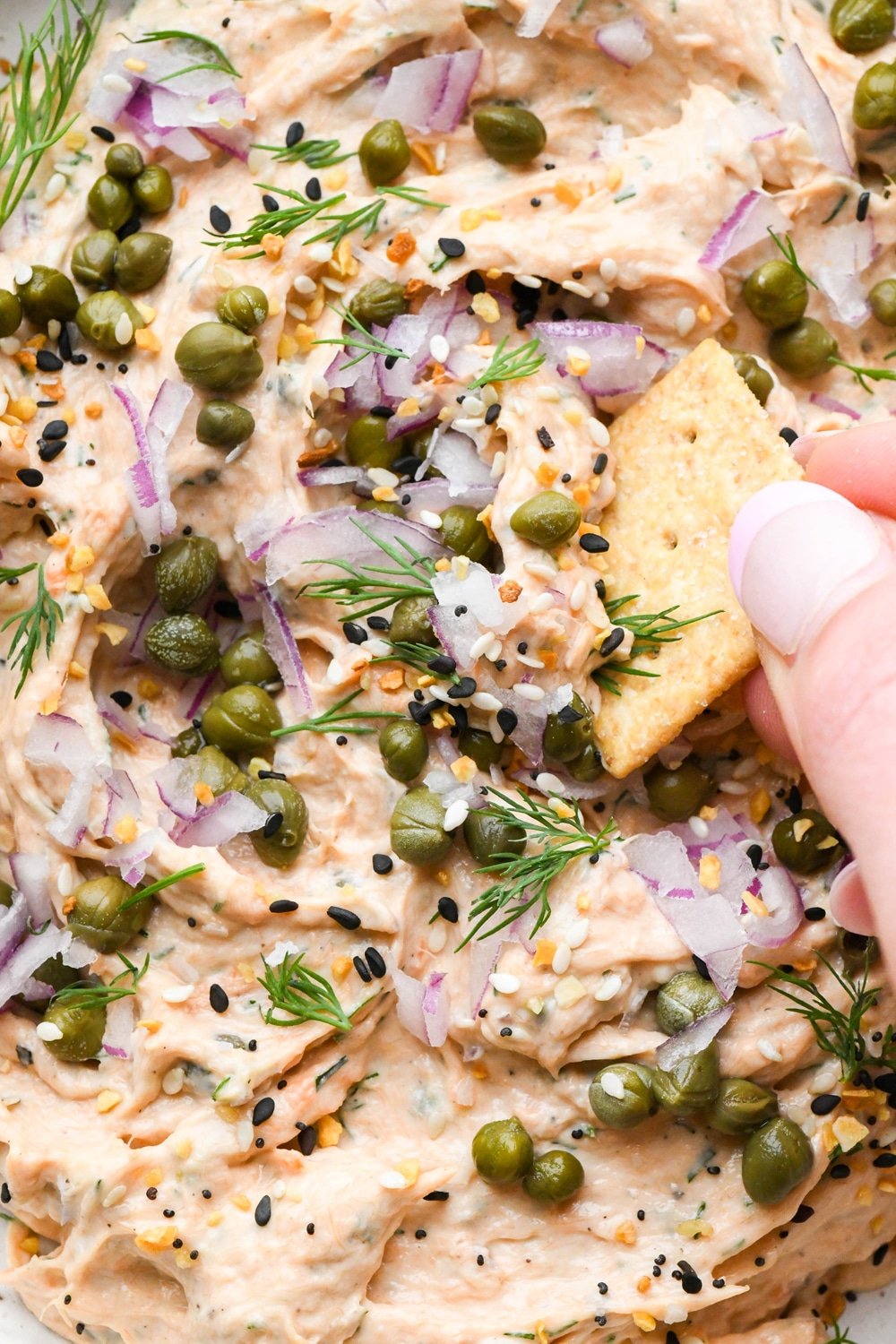 Dairy free smokes salmon dip in a small shallow ceramic speckled bowl, garnished with capers, fresh dill, everything but the bagel seasoning, and square gluten free crackers tucked into the dip and a hand dipping a cracker into the dip to lift it out.