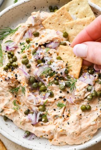 Dairy free smoked salmon dip in a small shallow ceramic speckled bowl, garnished with capers, fresh dill, everything but the bagel seasoning, and square gluten free crackers tucked into the dip and a hand dipping a cracker into the dip to lift it out.