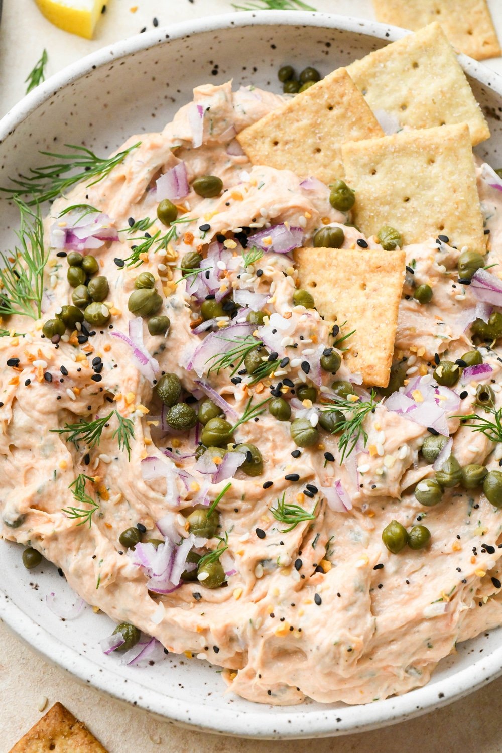 Dairy free smokes salmon dip in a small shallow ceramic speckled bowl, garnished with capers, fresh dill, everything but the bagel seasoning, and square gluten free crackers tucked into the dip.