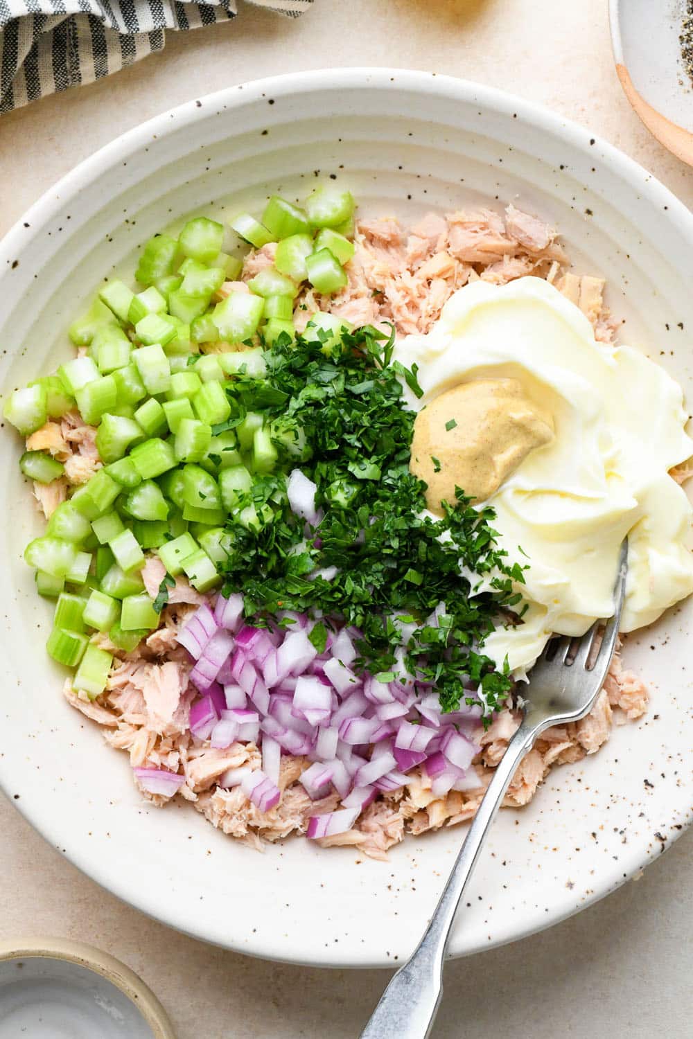 How to make tuna salad: Flaked tuna in a large bowl with diced celery, red onion, parsley, mayonnaise, and dijon mustard.
