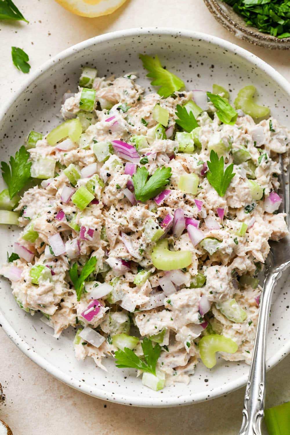 Tuna salad in a shallow ceramic bowl topped with some extra pieces of chopped celery, red onion, and parsley.