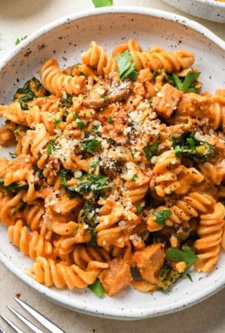 Creamy tomato sausage pasta in a shallow white speckled bowl, next to another bowl of pasta on a light cream background, and topped with dairy free parmesan, chili flakes, and a fresh herbs.