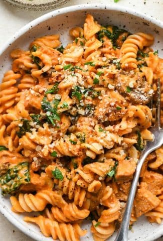 Creamy tomato sausage pasta in a shallow white speckled bowl, topped with dairy free parmesan, chili flakes, and a fresh herbs.