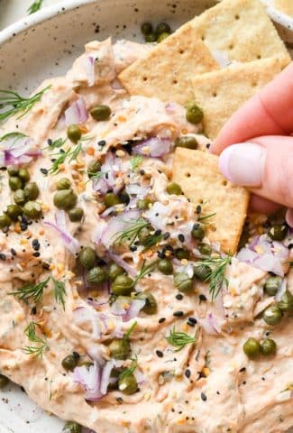 Dairy free smoked salmon dip in a small shallow ceramic speckled bowl, garnished with capers, fresh dill, everything but the bagel seasoning, and square gluten free crackers tucked into the dip and a hand dipping a cracker into the dip to lift it out.