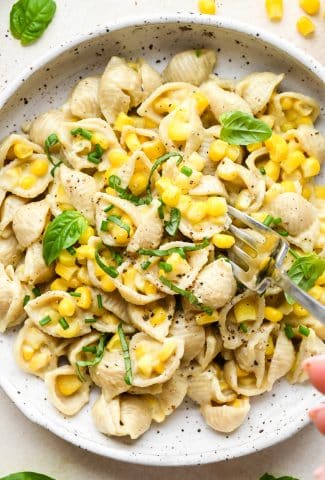 A shallow white speckled pottery bowl with creamy dairy free corn pasta topped with fresh basil and fresh chives, and a fork digging in for a bite.