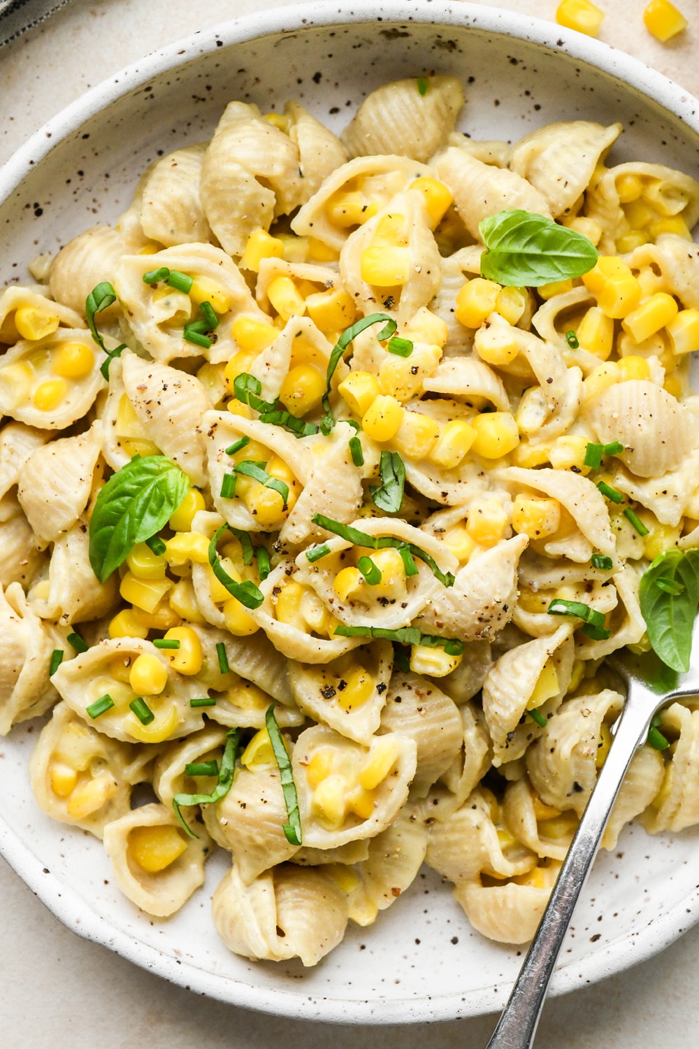 A shallow white speckled pottery bowl with creamy dairy free corn pasta topped with fresh basil and fresh chives.