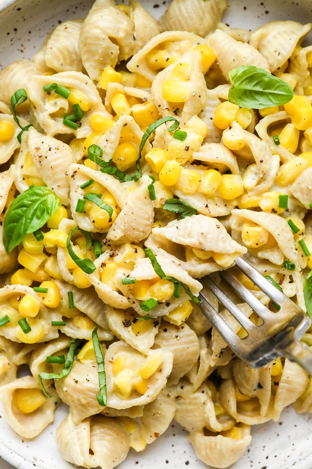 A close up image of a fork digging into a bowl of creamy dairy free corn pasta that is topped with fresh basil and cracked black pepper.