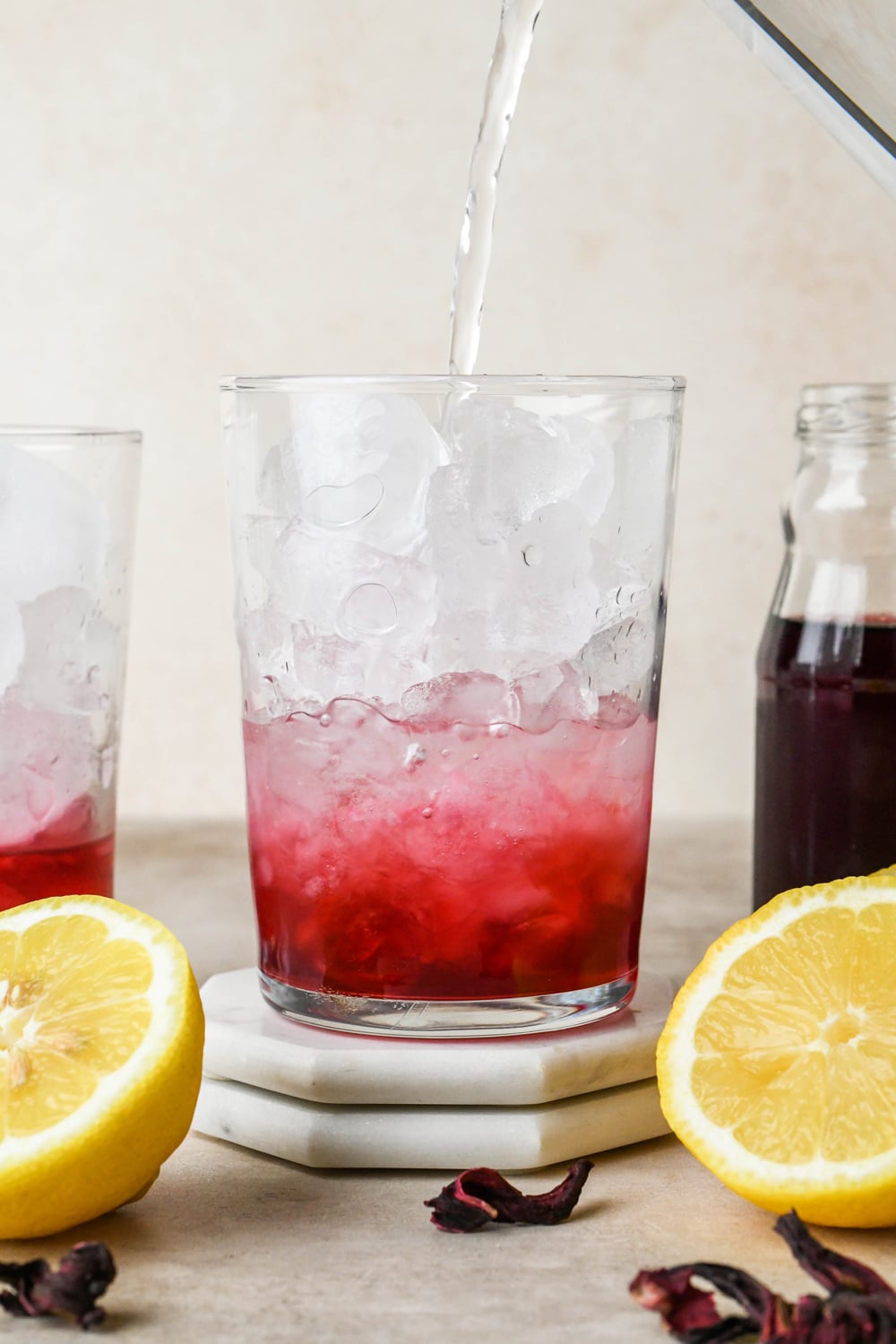 How to make hibiscus lemonade: Pouring water into a glass with hibiscus concentrate, lemon juice, and ice. 