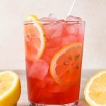 A straight on image of a single glass of hibiscus lemonade garnished with lemon wheels, surrounded by a few scattered dried hibiscus flowers and cut lemons.