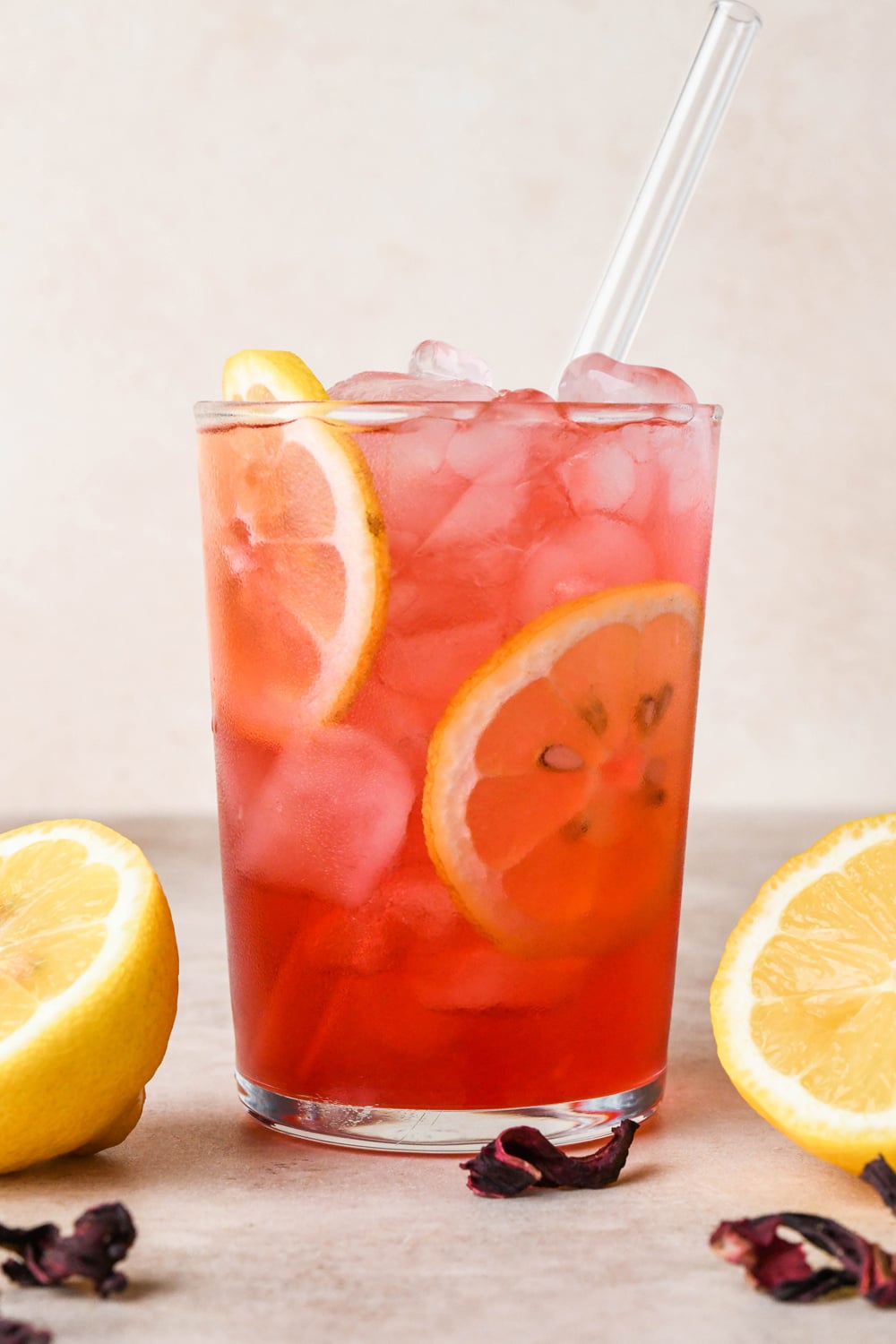 A straight on image of a single glass of hibiscus lemonade garnished with lemon wheels, surrounded by a few scattered dried hibiscus flowers and cut lemons.