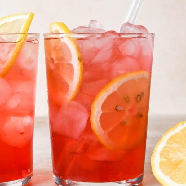 Two clear glasses of bright pink hibiscus lemonade sitting side by side against a creamy background, garnished with lemon wheels and dried hibiscus flowers.