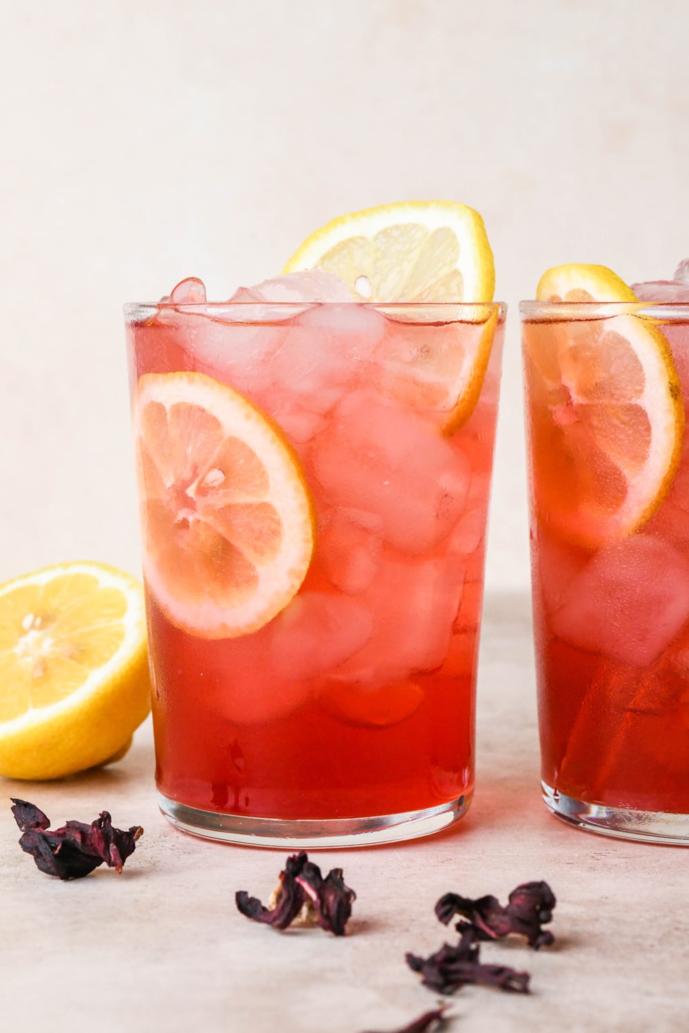 Two clear glasses of bright pink hibiscus lemonade sitting side by side against a creamy background, garnished with lemon wheels and dried hibiscus flowers, surrounded by some scattered dried hibiscus and cut lemons.