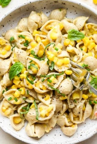 A shallow white speckled pottery bowl with creamy dairy free corn pasta topped with fresh basil and fresh chives, and a fork digging in for a bite.