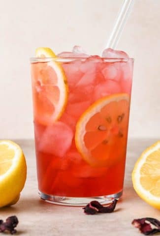 A straight on image of a single glass of hibiscus lemonade garnished with lemon wheels, surrounded by a few scattered dried hibiscus flowers and cut lemons.