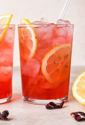 Two clear glasses of bright pink hibiscus lemonade sitting side by side against a creamy background, garnished with lemon wheels and dried hibiscus flowers.