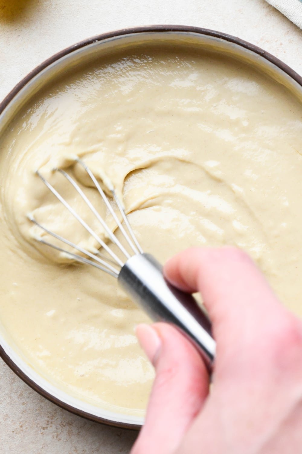A hand holding a small whisk, whisking creamy tahini dressing together in a shallow bowl.