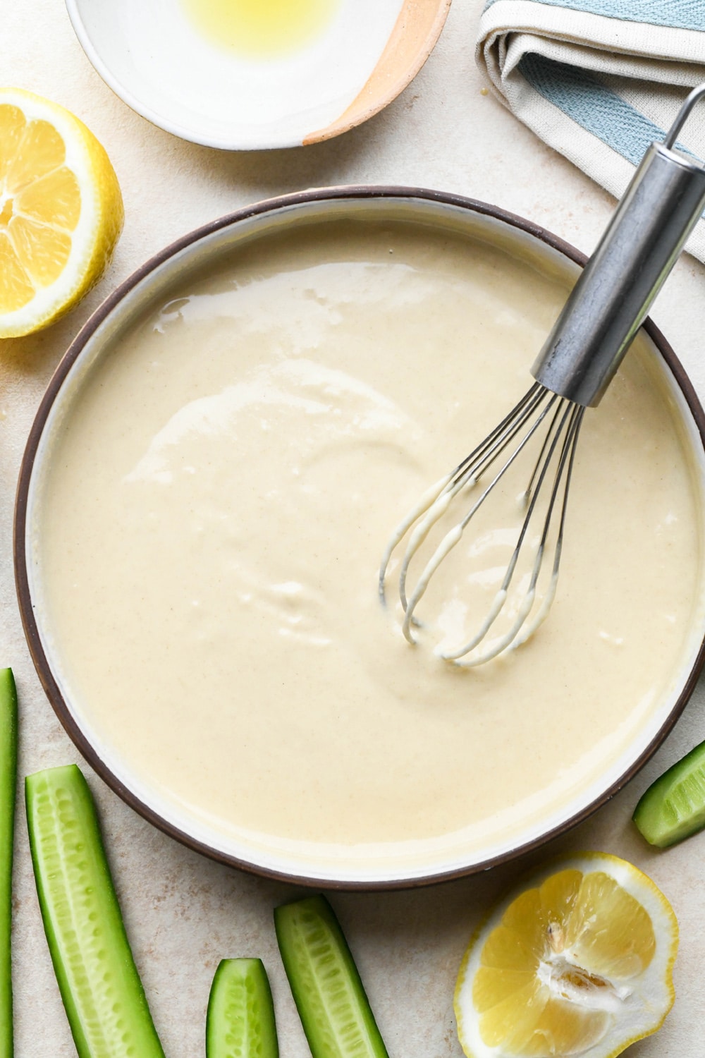 Creamy tahini dressing in a shallow bowl with a small whisk, surrounded by some cut cucumbers and lemons.