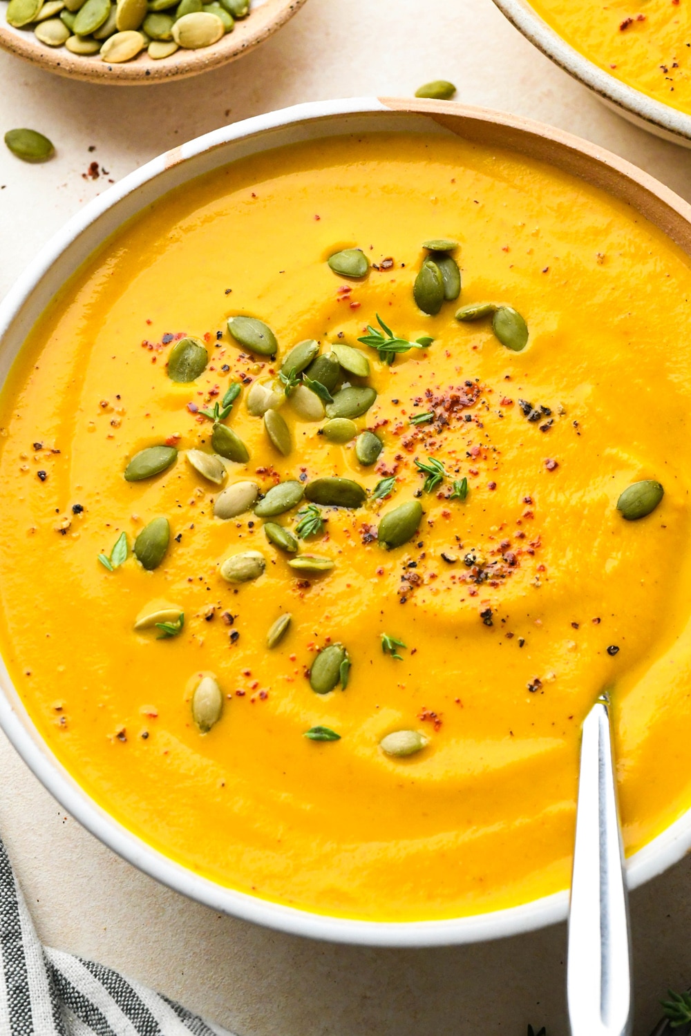 Bright orange dairy free pumpkin and carrot soup in a large soup bowl, garnished with pumpkin seeds, chili flakes, black pepper, and fresh thyme.