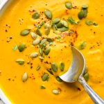 Bright orange dairy free pumpkin and carrot soup in a large soup bowl, with a spoon dipping into it, garnished with pumpkin seeds, chili flakes, black pepper, and fresh thyme.