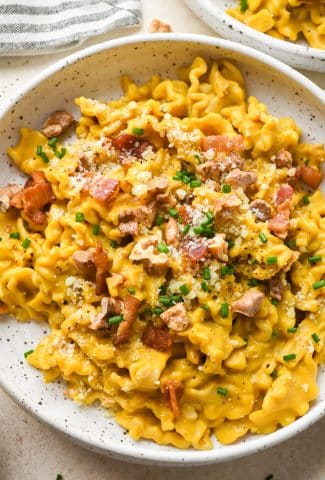 Creamy, dairy free butternut squash pasta with bacon, walnuts, and chives, in a shallow speckled ceramic bowl on a light cream background, next to a striped napkin and a second bowl of pasta.