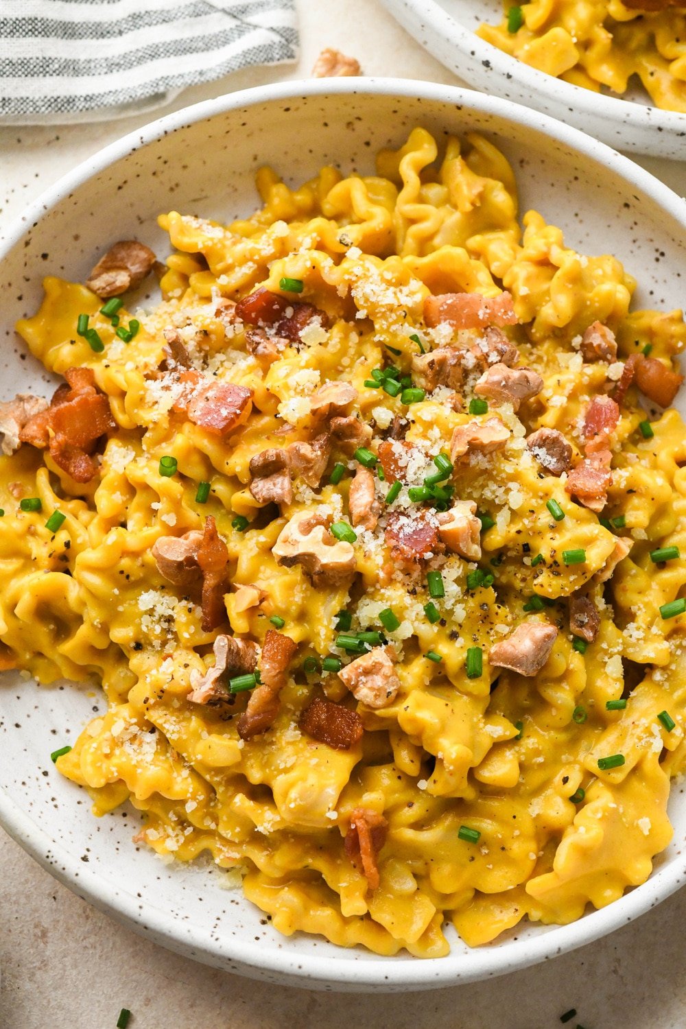 Creamy, dairy free butternut squash pasta with bacon, walnuts, and chives, in a shallow speckled ceramic bowl on a light cream background, next to a striped napkin and a second bowl of pasta.