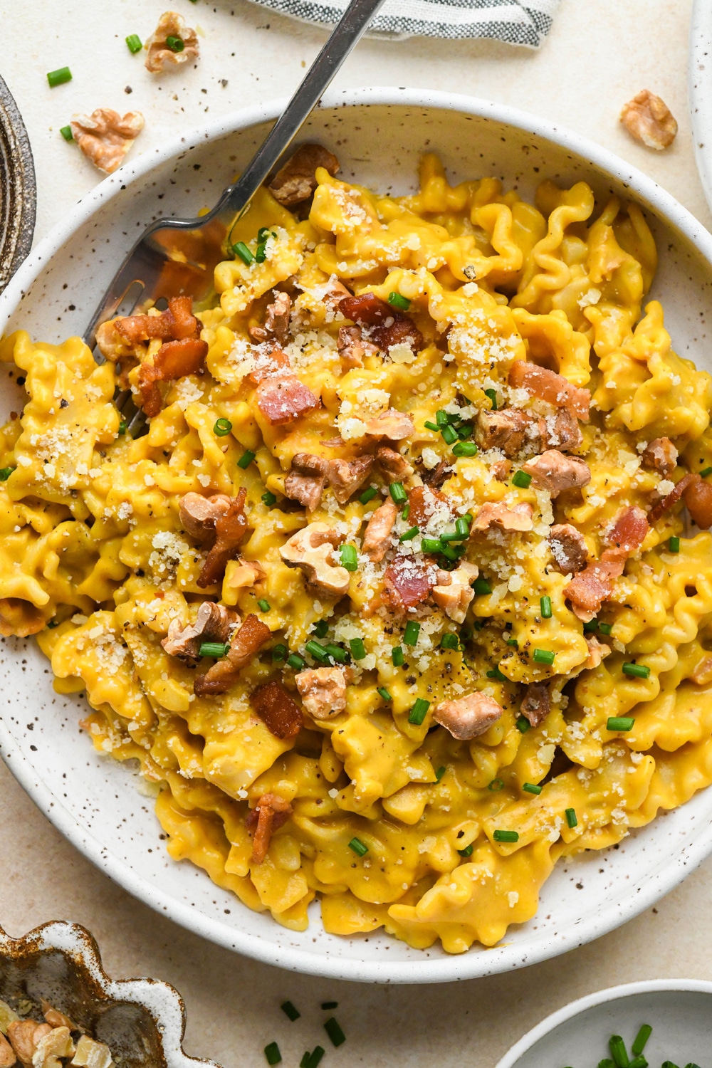 Creamy, dairy free butternut squash pasta with bacon, walnuts, and chives, in a shallow speckled ceramic bowl on a light cream background, with a fork angled into the bowl. 
