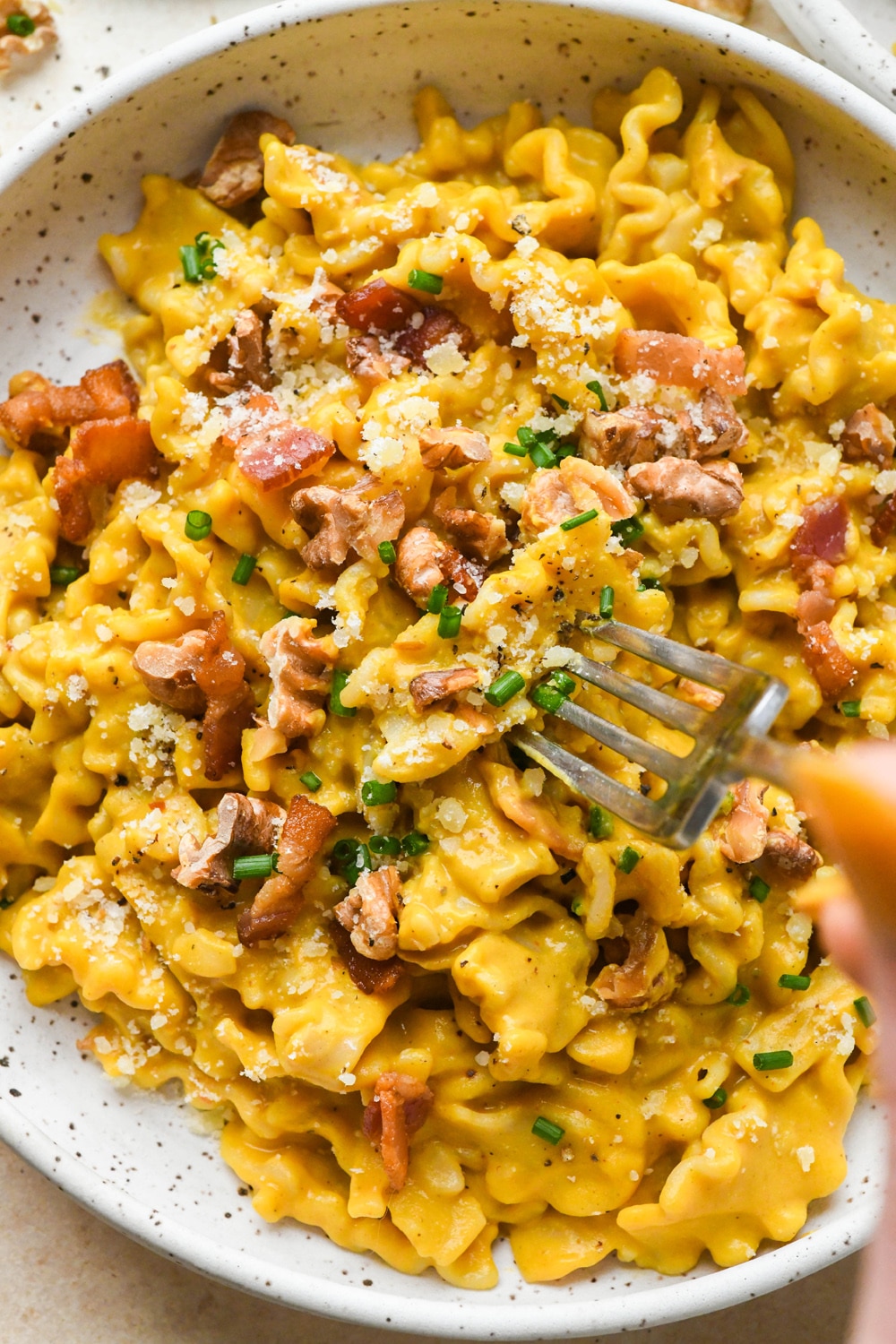 Creamy, dairy free butternut squash pasta with bacon, walnuts, and chives, in a shallow speckled ceramic bowl with a fork piercing a piece of pasta to show the creamy texture of the pasta sauce.