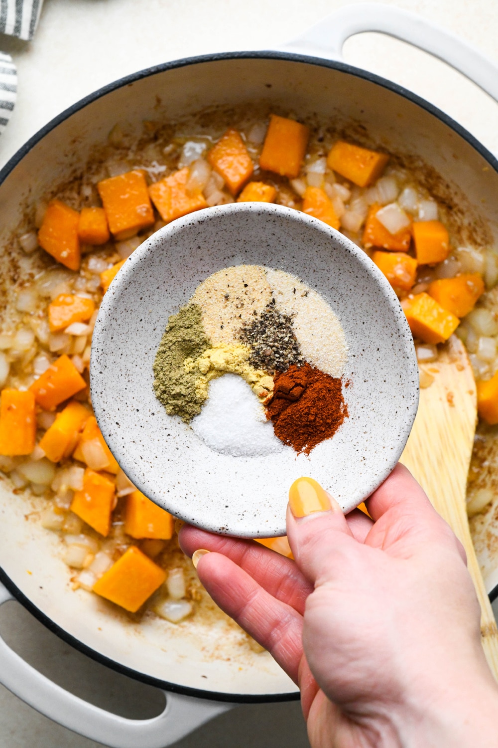 How to make Dairy Free Butternut Squash Pasta: Small dish of spices being held above the sautéed butternut squash, onions, and garlic to demonstrate adding it to the skillet. 