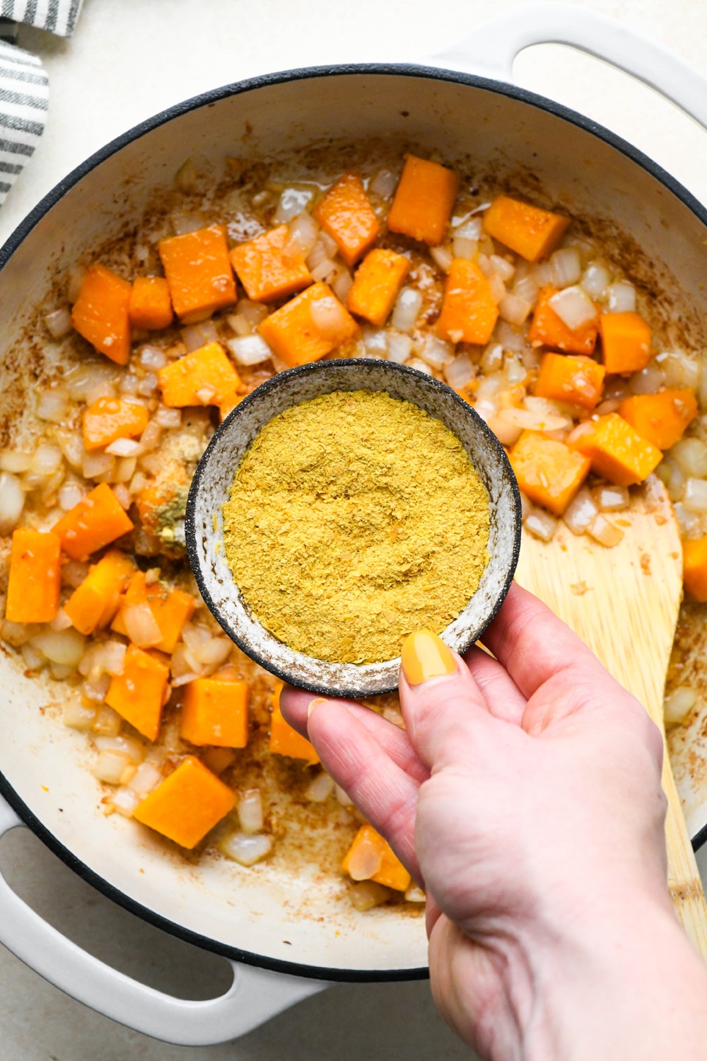 How to make Dairy Free Butternut Squash Pasta: Small dish of nutritional yeast being held above the sautéed butternut squash, onions, and garlic to demonstrate adding it to the skillet. 